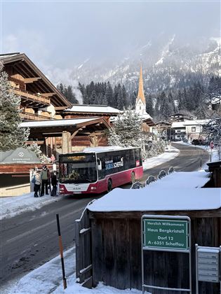 Een bus stopt in een besneeuwd dorp. Op de achtergrond zijn bergen en een kerk met een spitse toren te zien.
