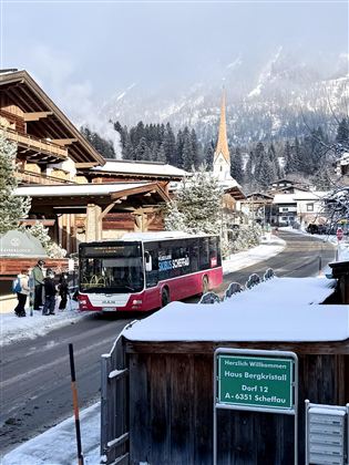 Ein Winterblick auf ein malerisches Dorf mit schneebedeckten Bergen. Ein Bus hält an einer Haltestelle, während Passagiere darauf warten.