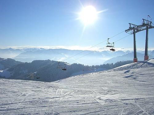 Eine wunderschöne Winterlandschaft mit schneebedeckten Bergen. Skilifte sind im Vordergrund, und die Sonne strahlt am klaren Himmel.