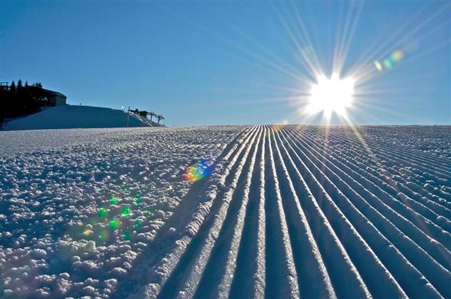 A snow-covered slope with perfectly groomed trails. The sun shines brightly in the clear blue sky.