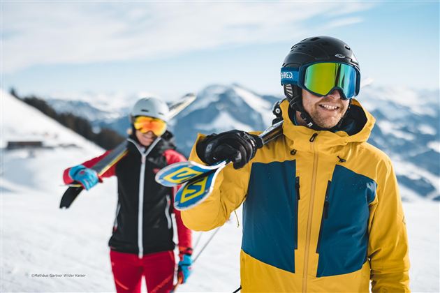 Two skiers are standing on a snow-covered slope in the mountains. The man is wearing a colorful ski suit and is smiling, while the woman in the background looks ready to ski.
