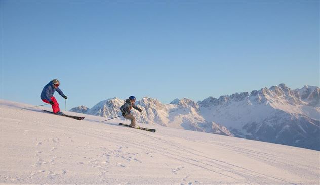 Zwei Skifahrer fahren fröhlich die schneebedeckte Piste hinunter. Im Hintergrund sind beeindruckende Berge und ein klarer, blauer Himmel zu sehen.