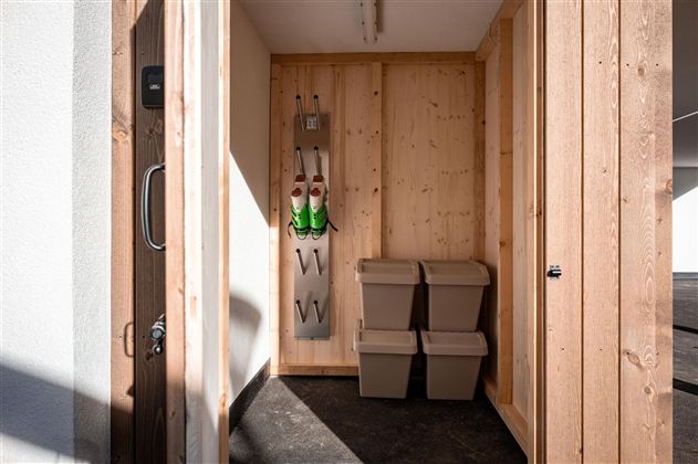 Ein kleiner Lagerraum mit Holzwänden und zwei Müllcontainern. An der Wand hängen zwei Grünglänzende Gegenstände und eine Aufbewahrungslösung.