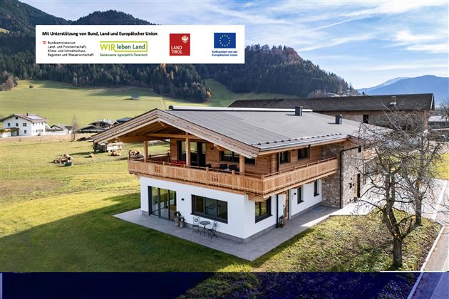 Ein modernes Holzhaus in einer ländlichen Umgebung mit grünen Wiesen und Bergen im Hintergrund. Der Himmel ist klar und die Architektur des Hauses ist ansprechend.