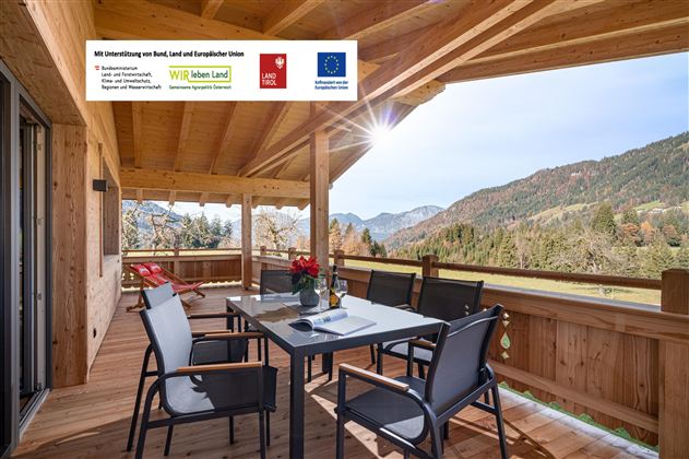 Eine einladende Terrasse mit einem Tisch und Stühlen, umgeben von malerischen Bergen. Die Holzstruktur verleiht der Umgebung eine warme und gemütliche Atmosphäre.