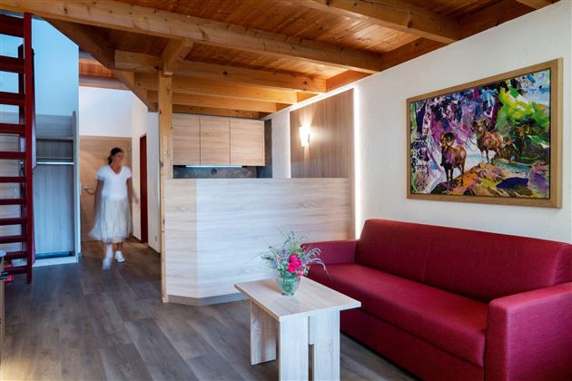 Ein modernes Wohnzimmer mit Holzdecke und rotem Sofa. Im Hintergrund ist eine Küchenzeile und ein farbenfrohes Gemälde zu sehen.