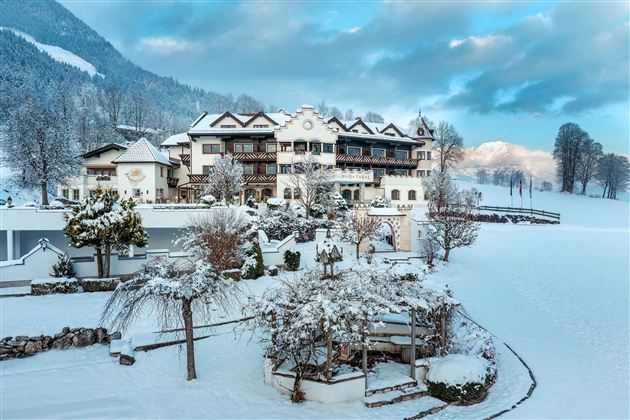Een idyllisch hotel in de sneeuw, omringd door besneeuwde bomen en bergen. De lucht is licht bewolkt en geeft de scène een rustige sfeer.