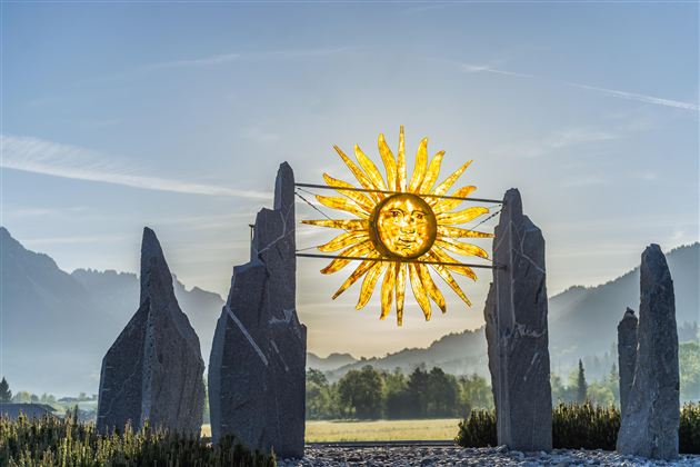 A sunny landscape featuring a large golden sun symbol placed between two stone sculptures. In the background, gentle mountains and a blue sky can be seen.