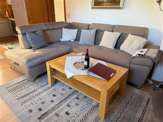 A cozy living room with a U-shaped sofa and many cushions. On the table, there is a bottle of wine, a glass, and a tablecloth.