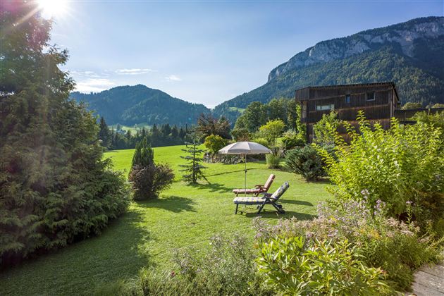 Ein schöner Garten mit Liegen und einem Sonnenschirm. Im Hintergrund sind grüne Berge und ein klarer Himmel zu sehen.