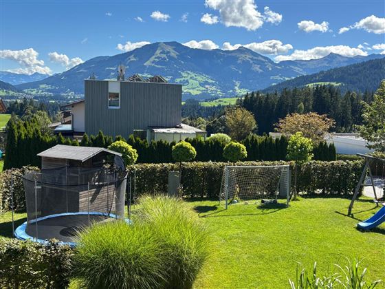 Ein schöner Garten mit einem Trampolin und Spielplatz. Im Hintergrund sieht man beeindruckende Berge und einen klaren Himmel.
