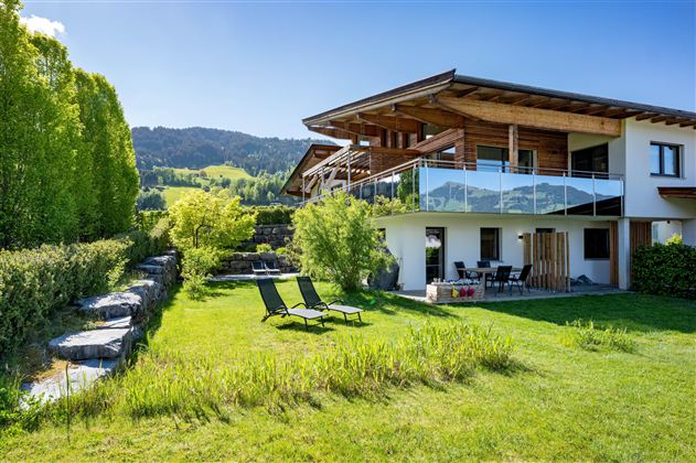 A modern house with a large garden and a terrace. The surroundings are green and hilly, ideal for relaxation.