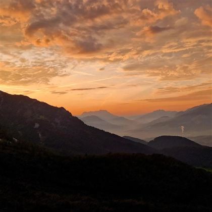 Een adembenemende zonsondergang boven de bergen, met oranje en paarse wolken. Het landschap toont zachte heuvels en een mystieke sfeer.