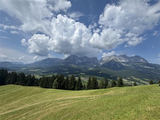 A picturesque landscape with green meadows and majestic mountains in the background. The sky is blue with white clouds.