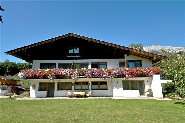 Ein schönes Holzhaus mit einem Balkon voller bunter Blumen. Der Garten ist grün und einladend, umgeben von Bäumen und Bergen.