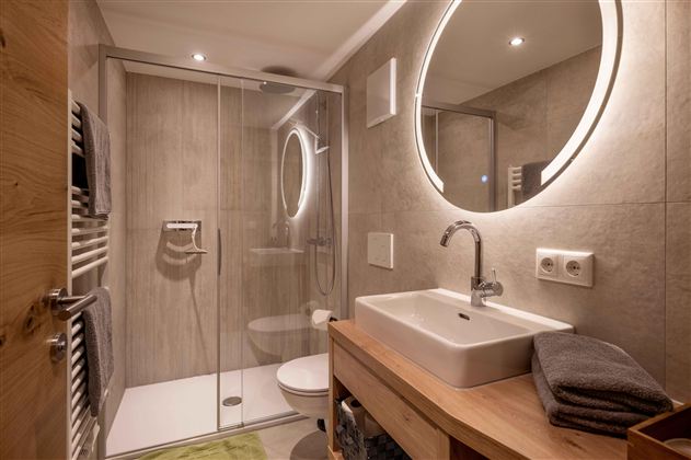 A modern bathroom with a glass shower and an elegant sink. The walls are kept in neutral colors, accompanied by a round, illuminated mirror surface.