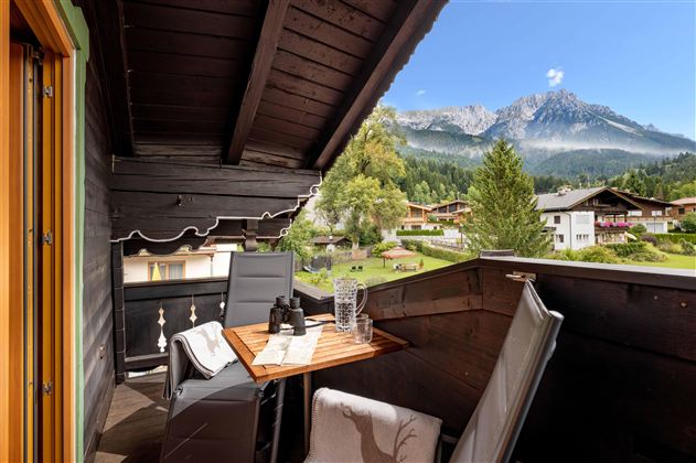 A cozy balcony with a table and chairs offers a beautiful view of the mountains and the surrounding landscape. The sun is shining, and it is a peaceful atmosphere.