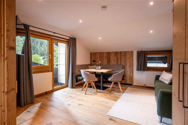 A cozy living space with a dining table, bright walls, and large windows. The room is decorated with modern furniture and wooden elements.