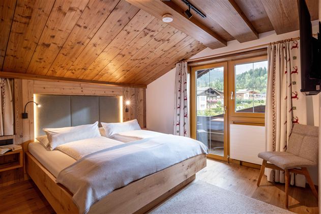 A cozy bedroom with a large double bed and wooden beam ceiling. Through the window, one looks out onto a green landscape.