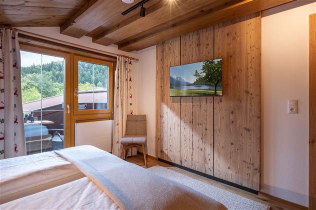 A bright bedroom with a wooden ceiling and a large window.  
A TV is mounted on the wall and a chair stands next to the bed.