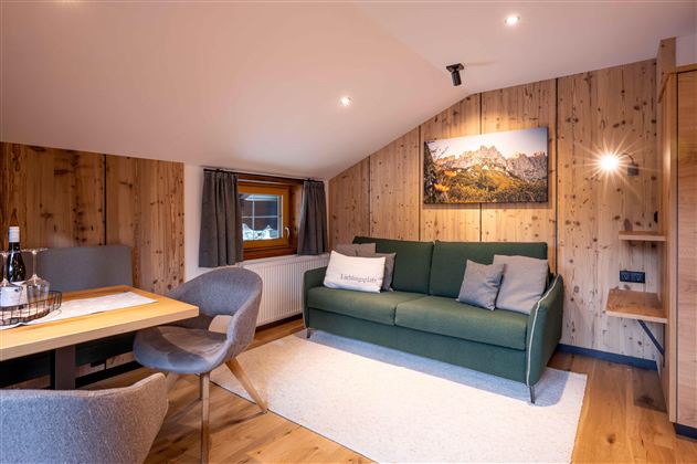 A cozy living room with a green sofa and a dining area. The walls are clad in wood and a picture of a mountain landscape hangs on the wall.