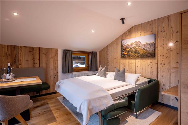A cozy bedroom with wooden paneling and a double bed. There is a small dining area and a window with a view of the mountains.