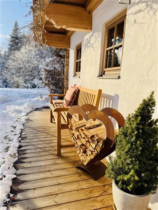 A cozy wooden bench with a heart motif, storing wood for the fireplace. The path is surrounded by fresh snow, and in the background, there are snow-covered trees.
