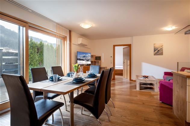 Eine modern eingerichtete Wohnung mit einem Essbereich und einem großen Fenster. Der Tisch ist gedeckt, und es gibt einen Fernseher sowie gemütliche Sitzgelegenheiten.