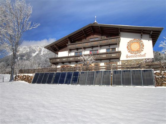Ein Fachwerkhaus im Schnee mit Solarpanels im Vordergrund. Im Hintergrund sind Berge und ein blauer Himmel zu sehen.
