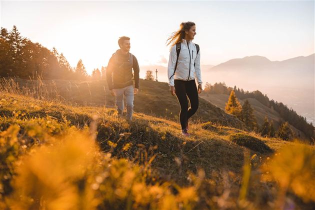 Two hikers enjoy nature at sunset. In the background, mountains and forests can be seen.