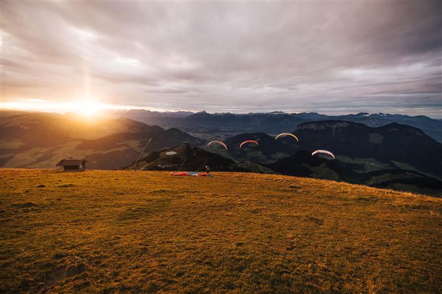A beautiful mountain landscape at sunset, with several paragliders flying over the hills. The clouds are soft, and the colors of the sky are warm and inviting.