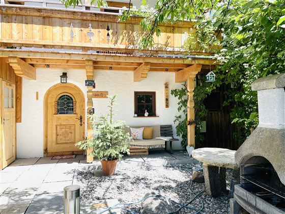 A cozy outdoor area with a wooden porch and plants. The entrance has a rustic door and there is a stone grill in the corner.