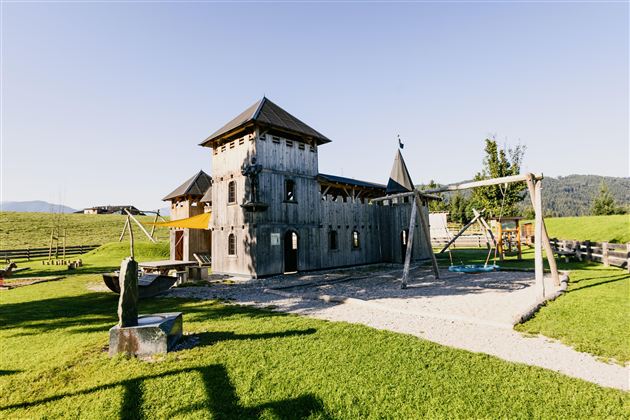 A playground in the shape of a castle with a large tower and swings. The surroundings consist of green grass and a clear blue sky.