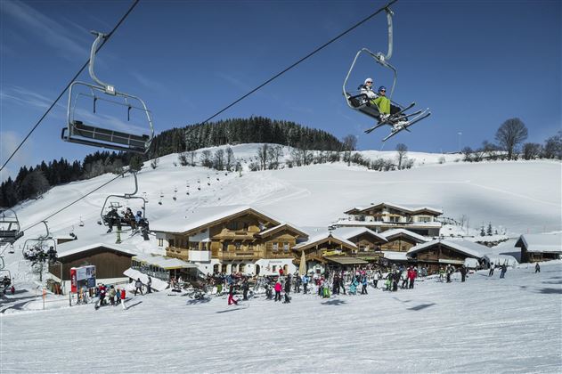 Ein Skigebiet mit schneebedeckten Bergen und einer Skilift-Anlage. Im Vordergrund sind zahlreiche Skifahrer und eine gemütliche Berghütte zu sehen.