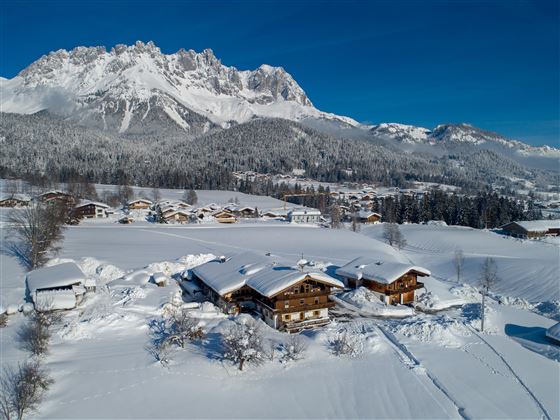 Eine winterliche Landschaft mit schneebedeckten Hütten und majestätischen Bergen im Hintergrund. Der klare blaue Himmel hebt die ruhige Atmosphäre der Szene hervor.
