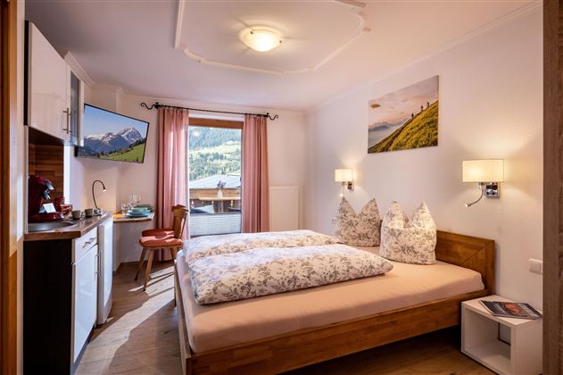 Ein gemütliches Schlafzimmer mit einem Doppelbett und dekorativen Kissen. Es gibt eine kleine Küchenzeile und Balkon mit Blick auf die Berge.