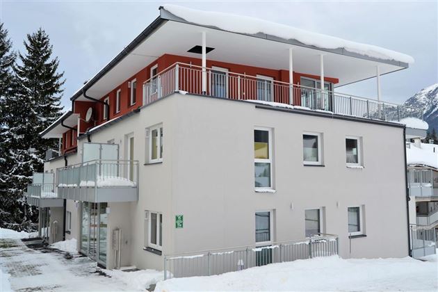 A modern building in the snow with white walls and red accents. The balcony is decorated with a snow overhang and there are some trees nearby.