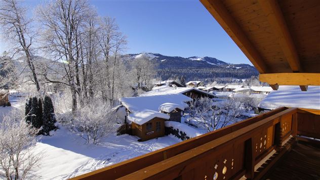 Eine winterliche Landschaft mit schneebedeckten Dächern und Bäumen. Im Hintergrund sind die Berge und ein klarer blauer Himmel zu sehen.