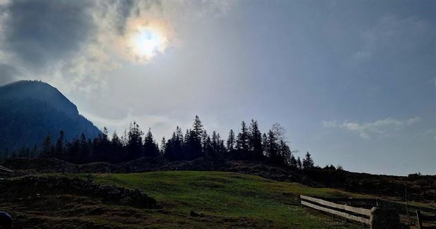 Een landschap met een zachte heuvel, omgeven door bomen en een bergachtige horizon. De lucht is bewolkt, en de zon schijnt door de wolken.
