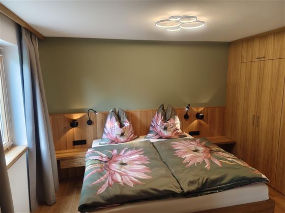 Een knusse slaapkamer met een tweepersoonsbed en bloemen decoratie op het beddengoed. De muren zijn in zachte kleuren gehouden, en er zijn moderne lampen naast het bed.