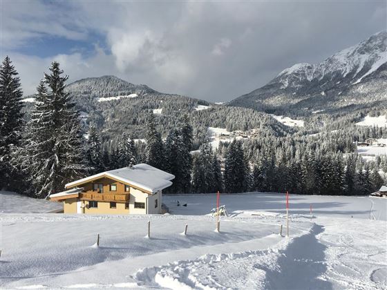 A snow-covered landscape with a cozy wooden house. In the background, snow-capped mountains and tall fir trees can be seen.