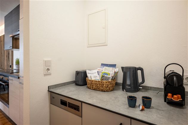 Eine moderne Küche mit einem grauen Arbeitsbereich. Auf der Theke stehen ein Wasserkocher, eine Kaffeekapselmaschine und eine Korb mit Zeitschriften.