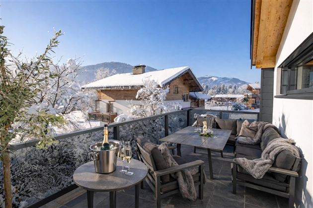 Eine gemütliche Terrasse mit einem Tisch und Stühlen, umgeben von schneebedeckten Bergen. Im Hintergrund sind traditionelle Holzhäuser und blauer Himmel zu sehen.