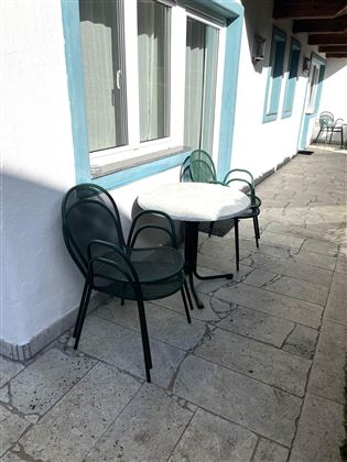 Ein kleiner Tisch mit zwei Stühlen steht auf einem gepflasterten Gehweg. Die Wand ist weiß mit blauen Fensterrahmen.