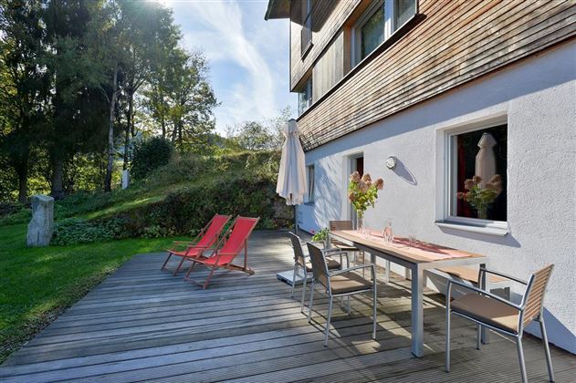 A cozy terrace with a wooden table, chairs, and red lounge chairs. In the background, green trees and a clear sky can be seen.