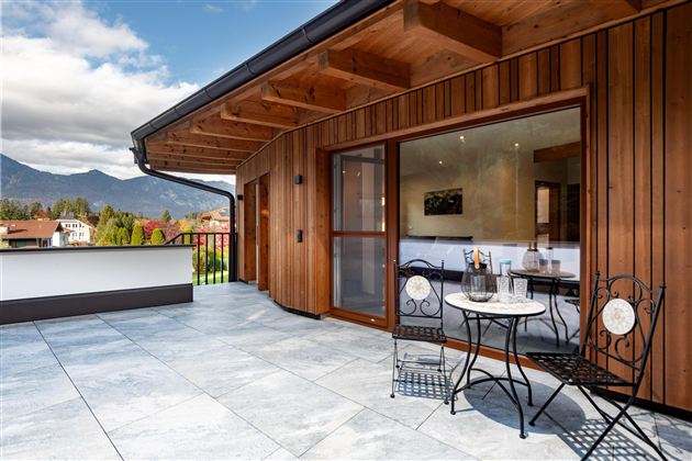 Eine moderne Terrasse mit Holzverkleidung und Blick auf die Berge. Ein kleiner Tisch mit Stühlen lädt zum Entspannen ein.