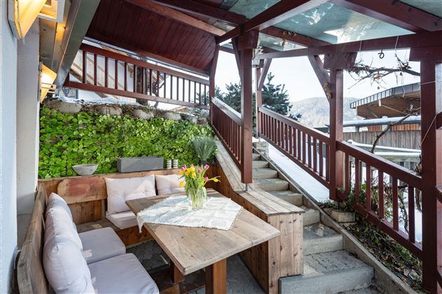 Eine gemütliche Terrasse mit Holzmöbeln und einer Vase mit Blumen. Im Hintergrund sind Stufen und eine grüne Wand zu sehen.