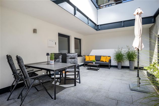 Ein moderner Innenhof mit einer gemütlichen Sitzgruppe und einem Tisch. Die Pflanzen und der Sonnenschirm sorgen für eine einladende Atmosphäre.