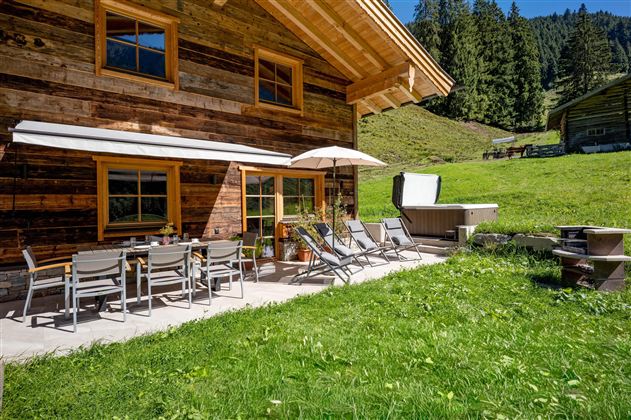 Eine gemütliche Terrasse mit einem Tisch und Liegestühlen, umgeben von grünem Gras und Bäumen. Im Hintergrund ist ein Holzhaus in einer malerischen Berglandschaft zu sehen. 
Chalet Heualm - Kelchsau Kitzbüheler Alpen