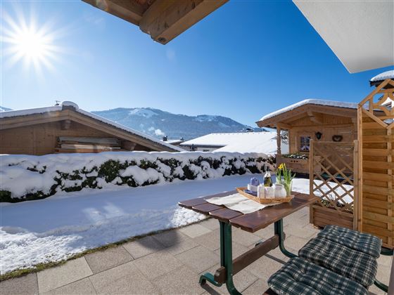 A sunny winter morning with snow-covered roofs and mountains in the background. On a terrace, there is a table with some drinks and a blooming plant arrangement.
